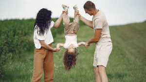 A joyful family enjoys outdoor play, lifting their child upside down in a green field.